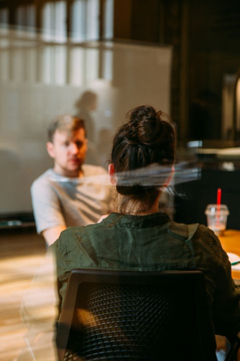 Two people talking across a table in a sunlit café, with coffee cups visible.