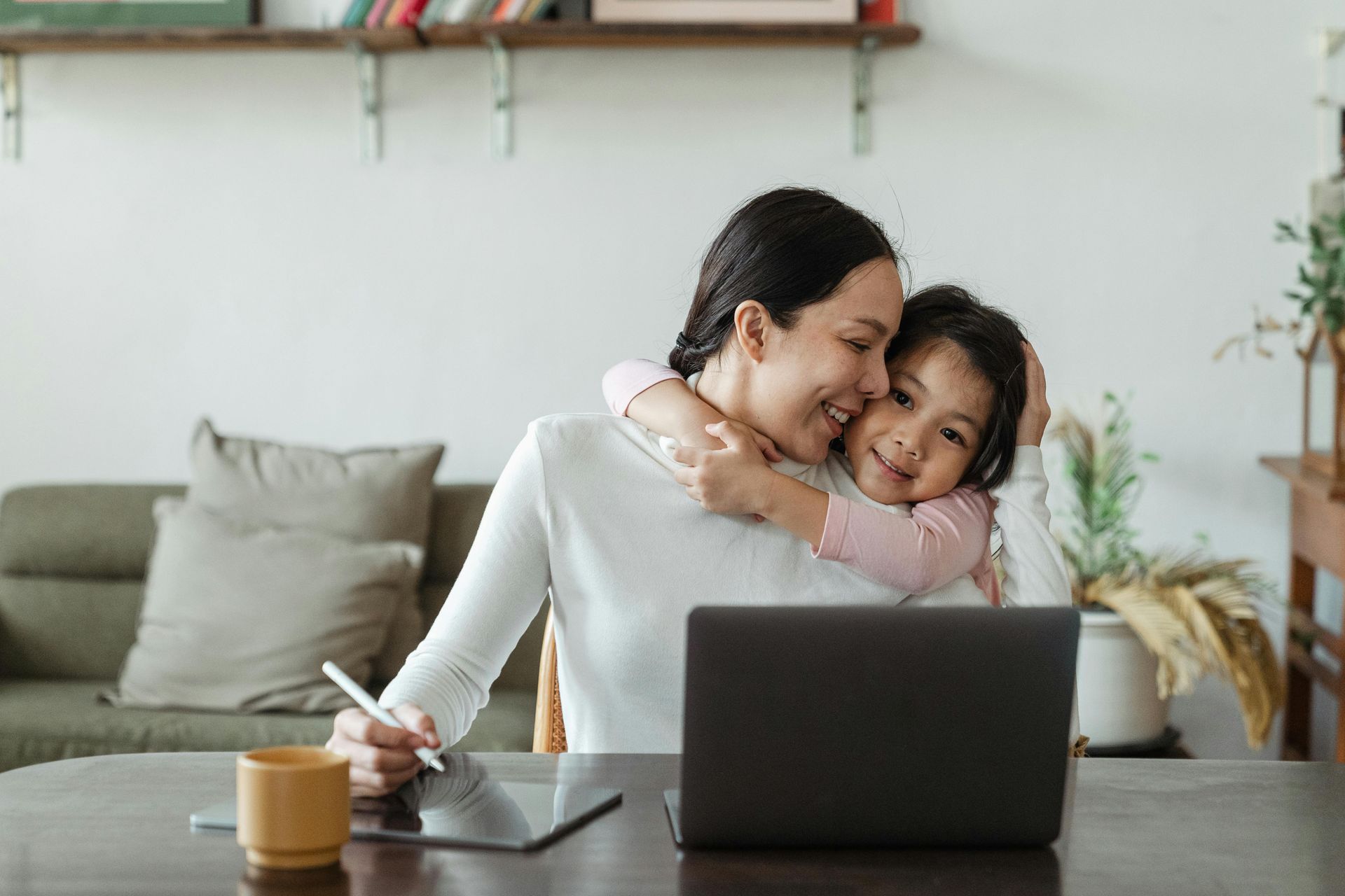 Woman at a table with a laptop and tablet is hugged by a smiling child. They are in a bright, homey setting.