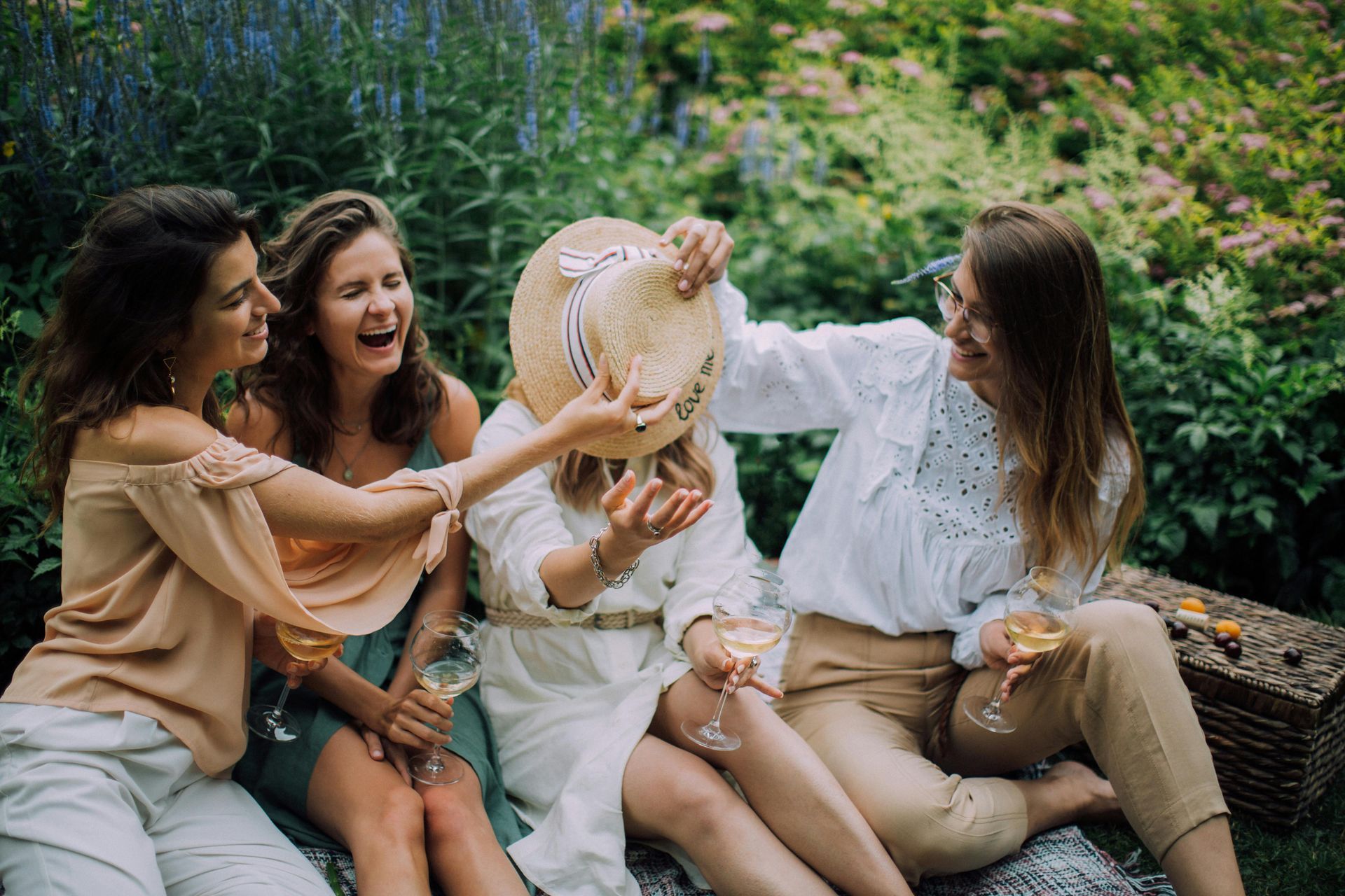 Four women laughing together at a picnic in a garden. One woman is playfully covering another's face with a straw hat.