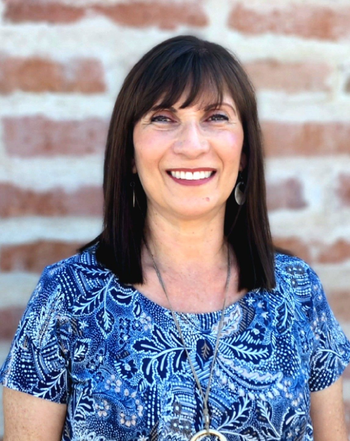 A woman in a blue shirt is smiling in front of a brick wall.