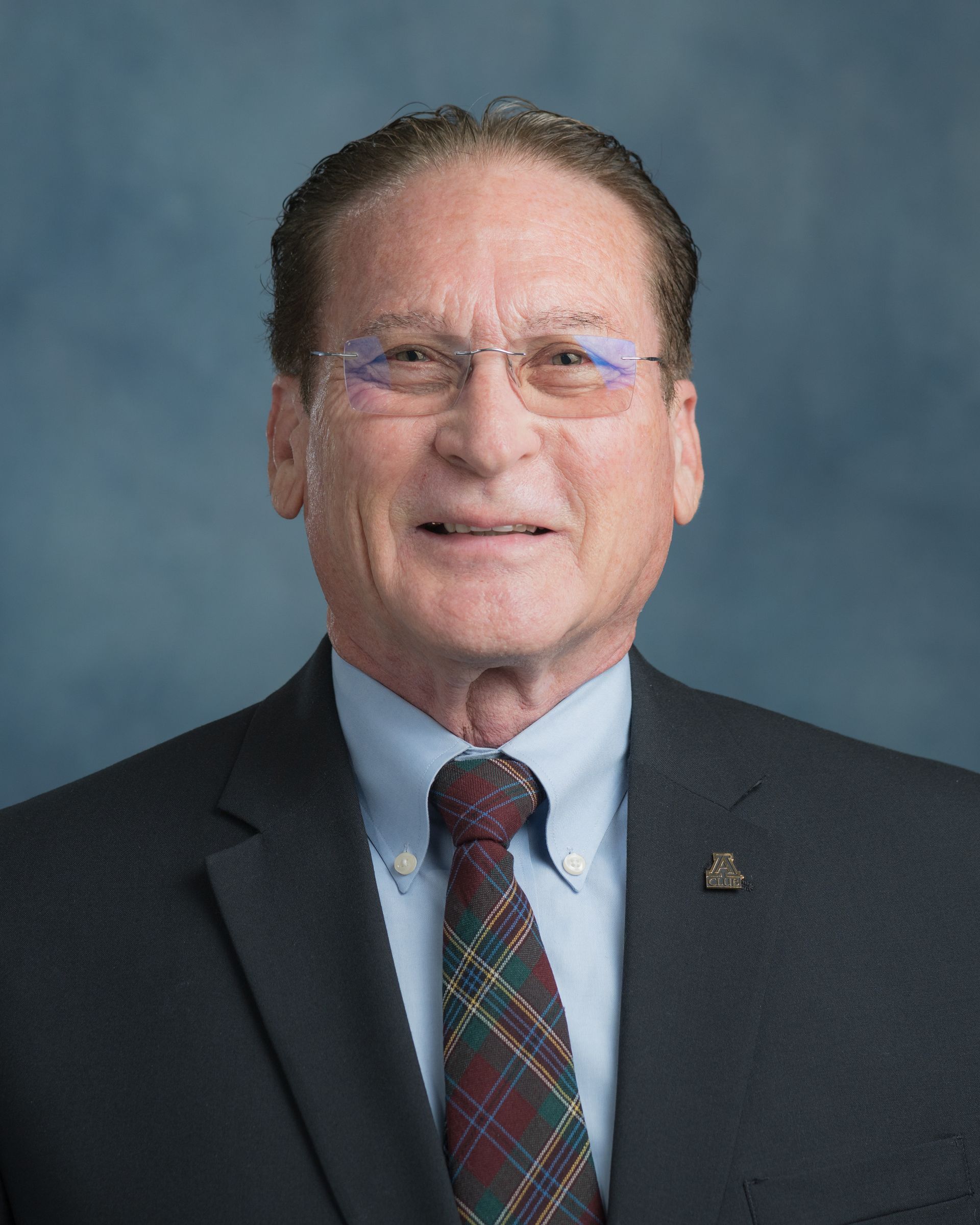 Man in suit and tie, wearing glasses, smiling, with blue and brown background.