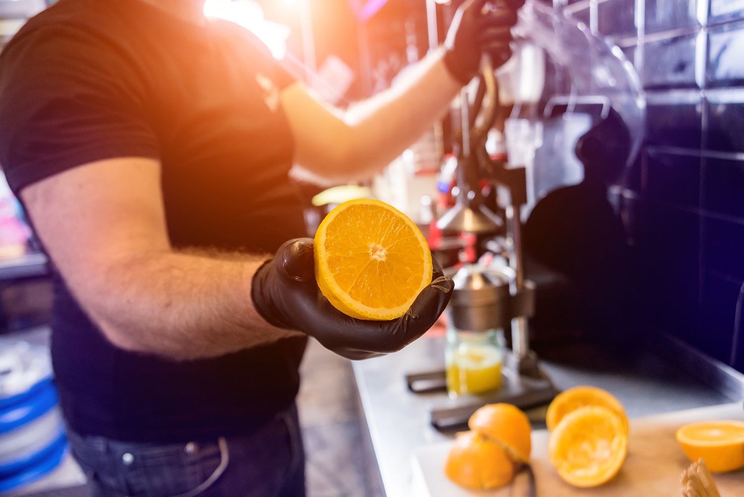 A gloved man holds an orange half while using a metal juicer inside a kitchen.