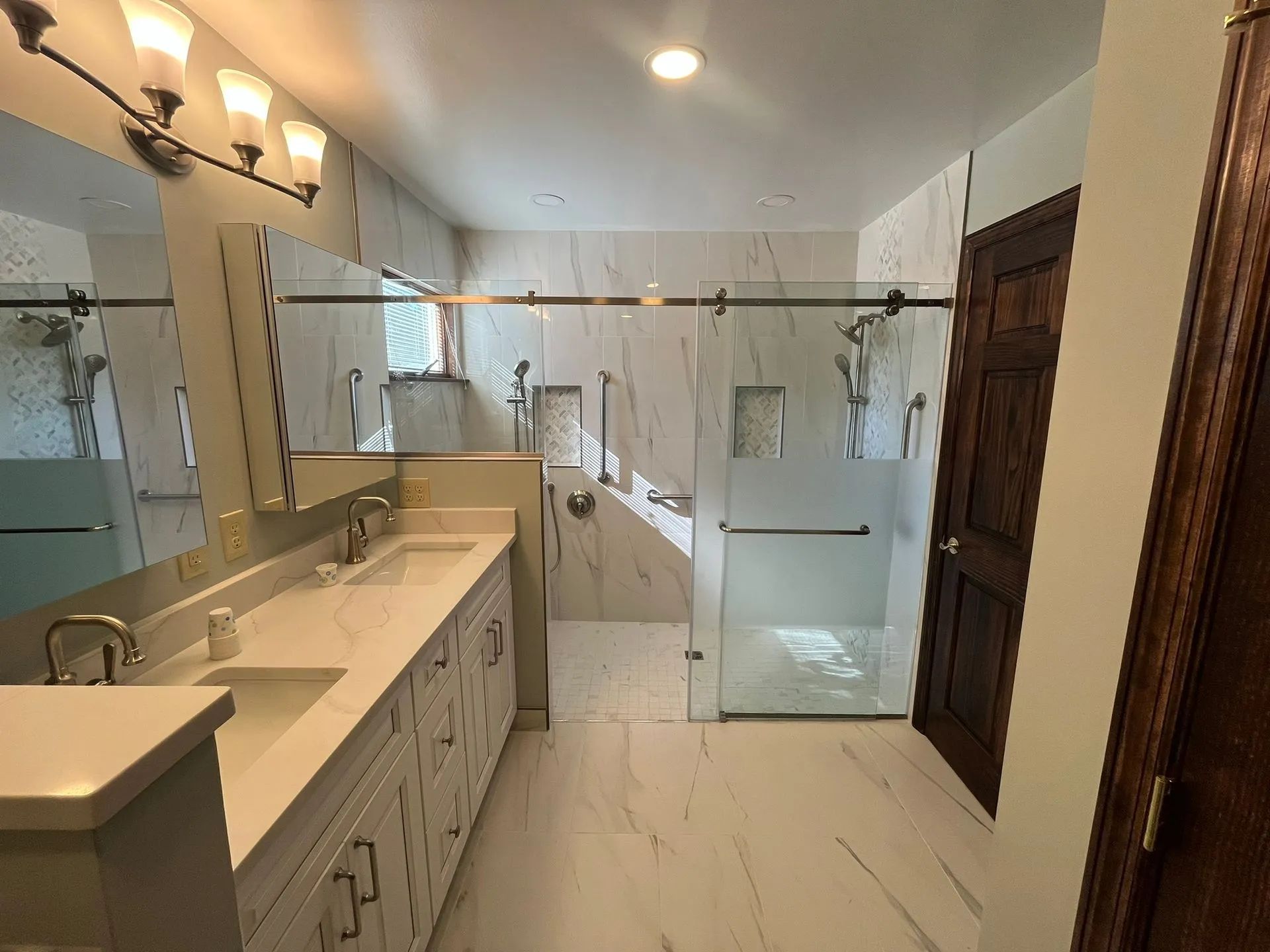 Bright bathroom with double vanity, mirrors, glass shower, and wood door on the right.