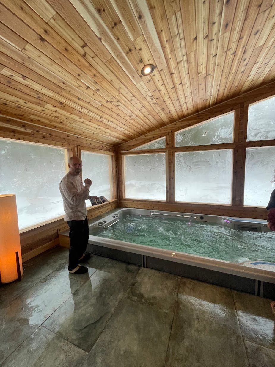 A man is standing next to a large hot tub in a room with a wooden ceiling.