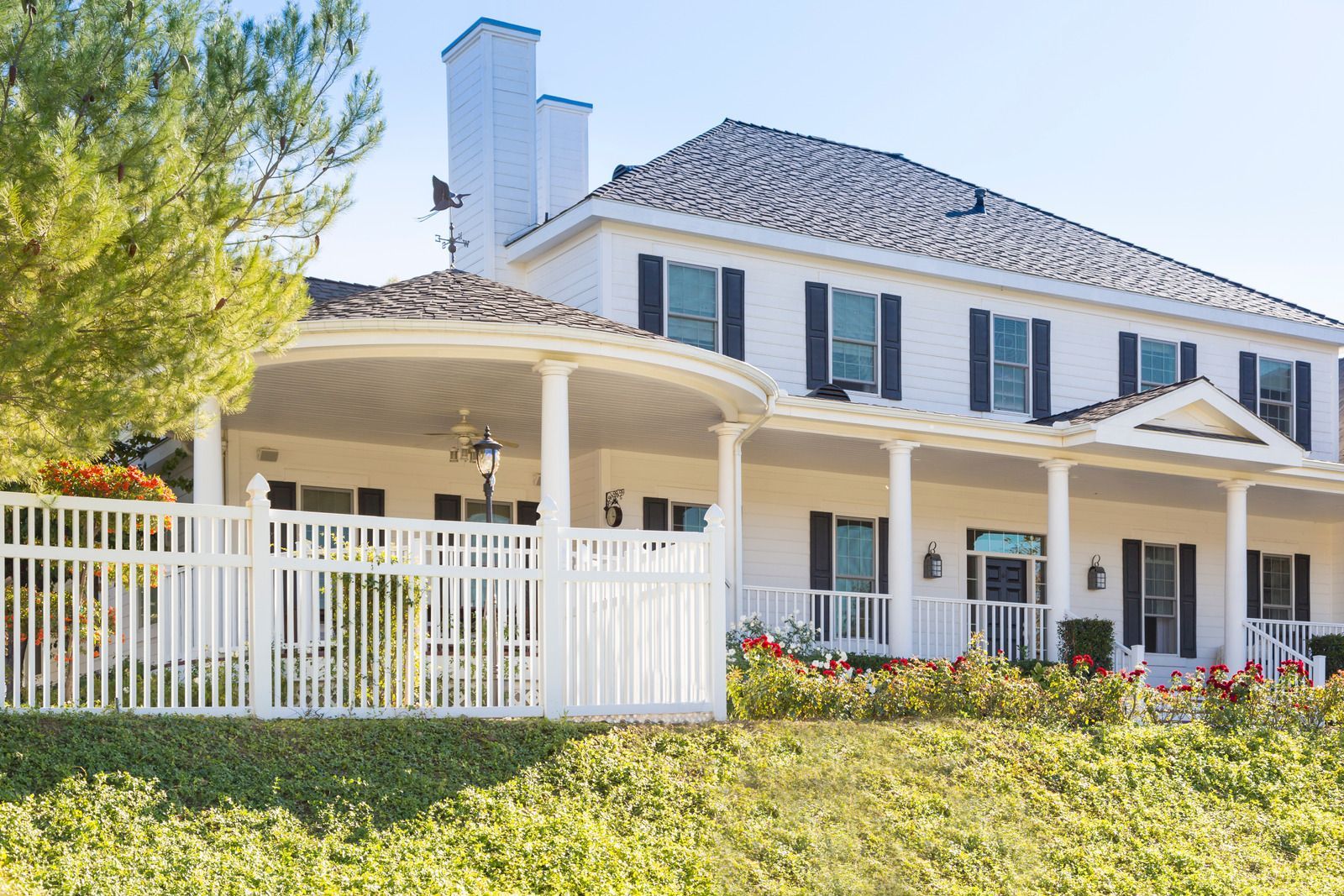 White two-story house with black shutters, a covered porch, and a white picket fence, set in a sunny, green landscape.