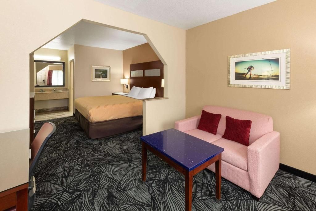 Hotel suite with bed, sofa, table, and bathroom, beige walls, dark carpet.