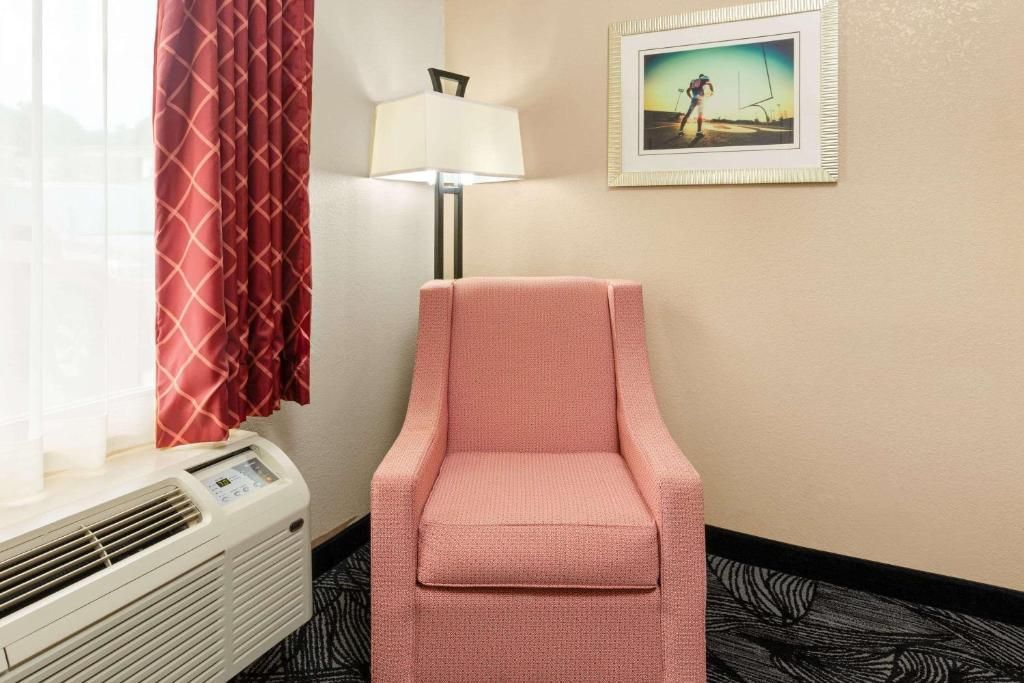 Pink upholstered armchair, lamp, and framed art in a hotel room, next to a window with red curtains and an air conditioner.