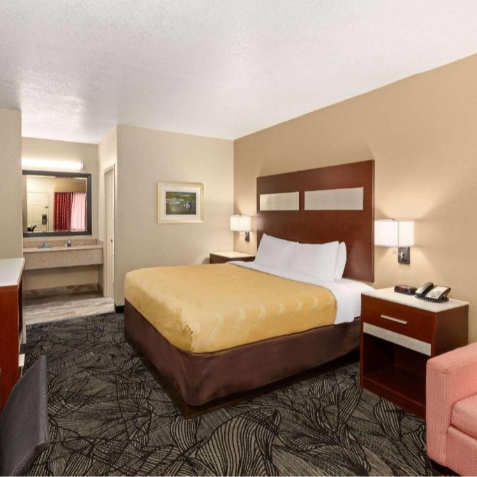 Hotel room with a queen bed, nightstand, and bathroom visible. Carpeted floor, beige walls.