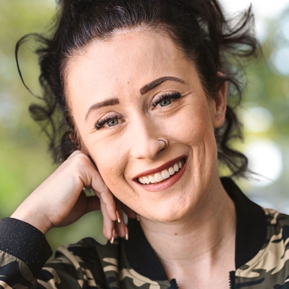 SmileArt patient with dark hair smiles, resting head on hand. Wearing camouflage jacket, outdoor setting.