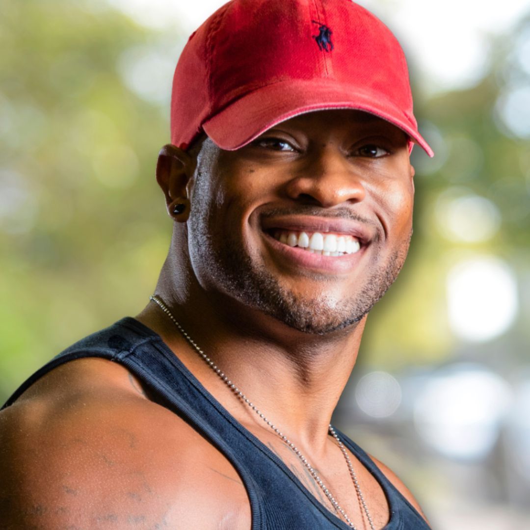 SmileArt patient smiling, wearing a red cap, tank top, and necklace. Outdoors, blurred background.