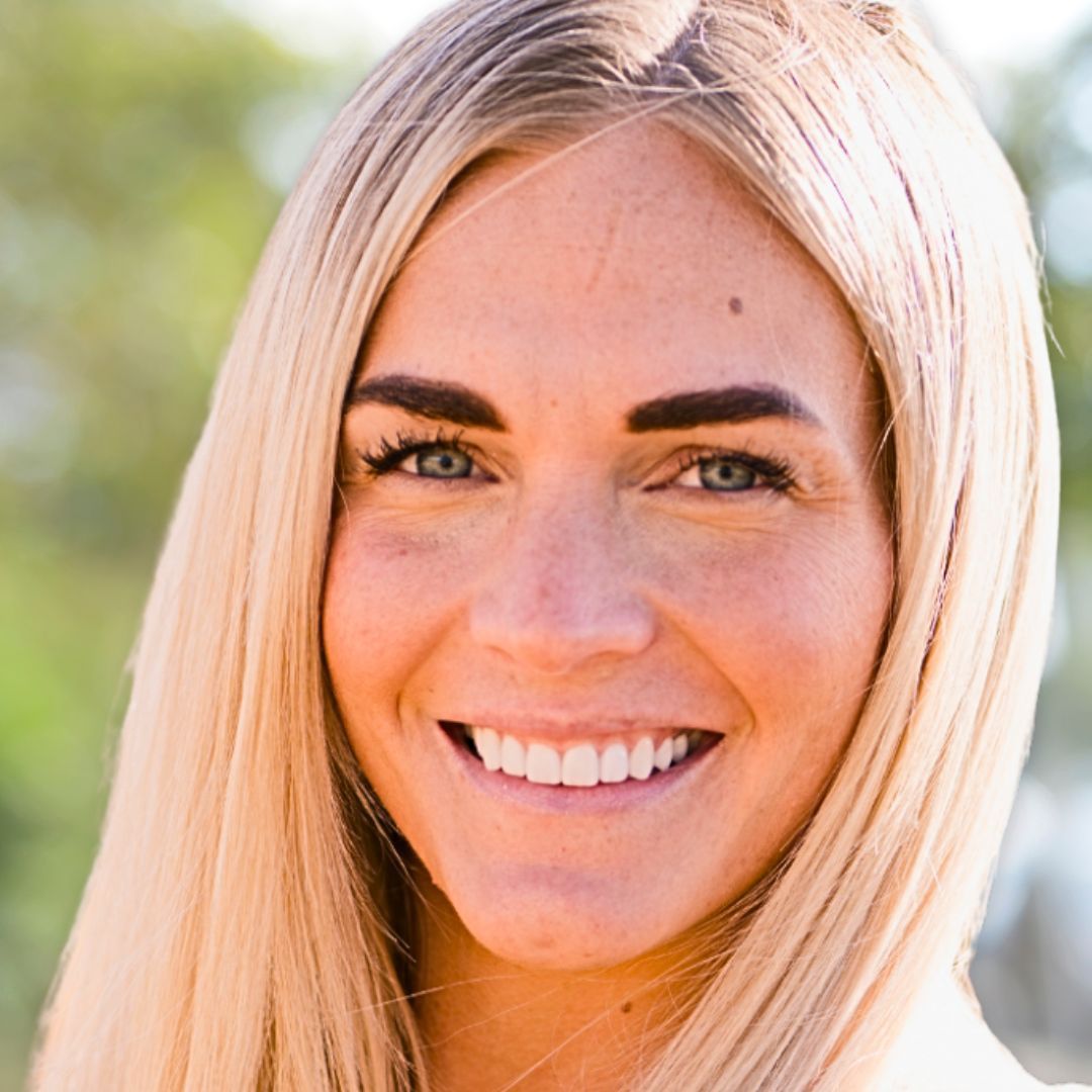 SmileArt patient with long blonde hair, smiling, looking directly at the viewer. Blurred outdoor background.