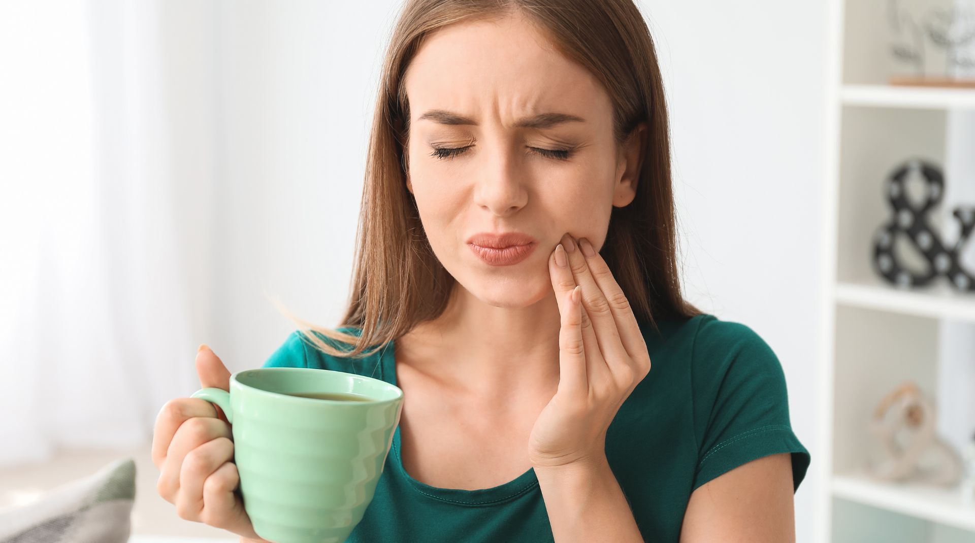 Woman holding a green mug, grimacing and touching her cheek, likely experiencing tooth pain.