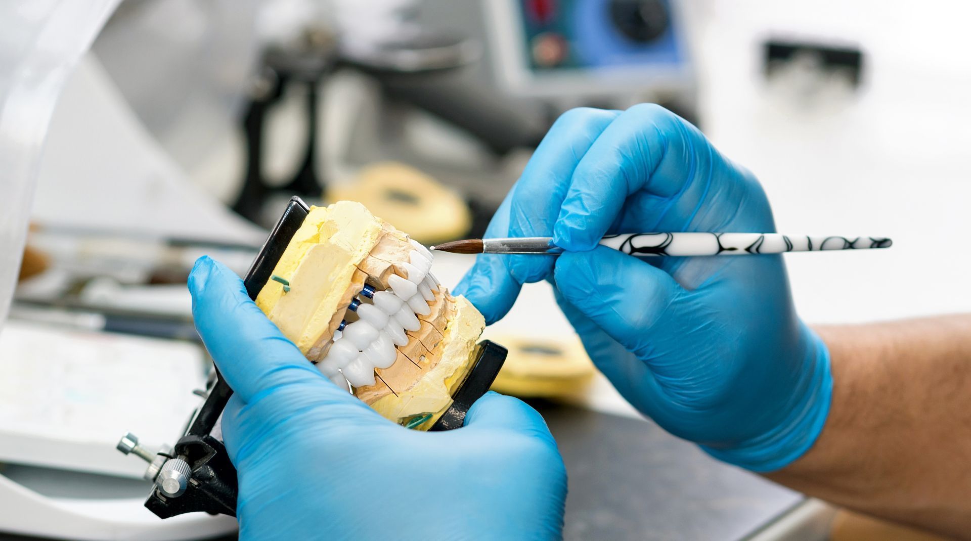 Dentist in blue gloves painting teeth model with small brush in a lab setting.