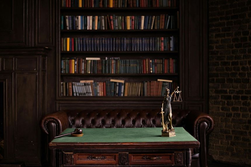 Dim study with a green-topped wooden desk, leather chair, bookshelves, and a small statue on the desk.