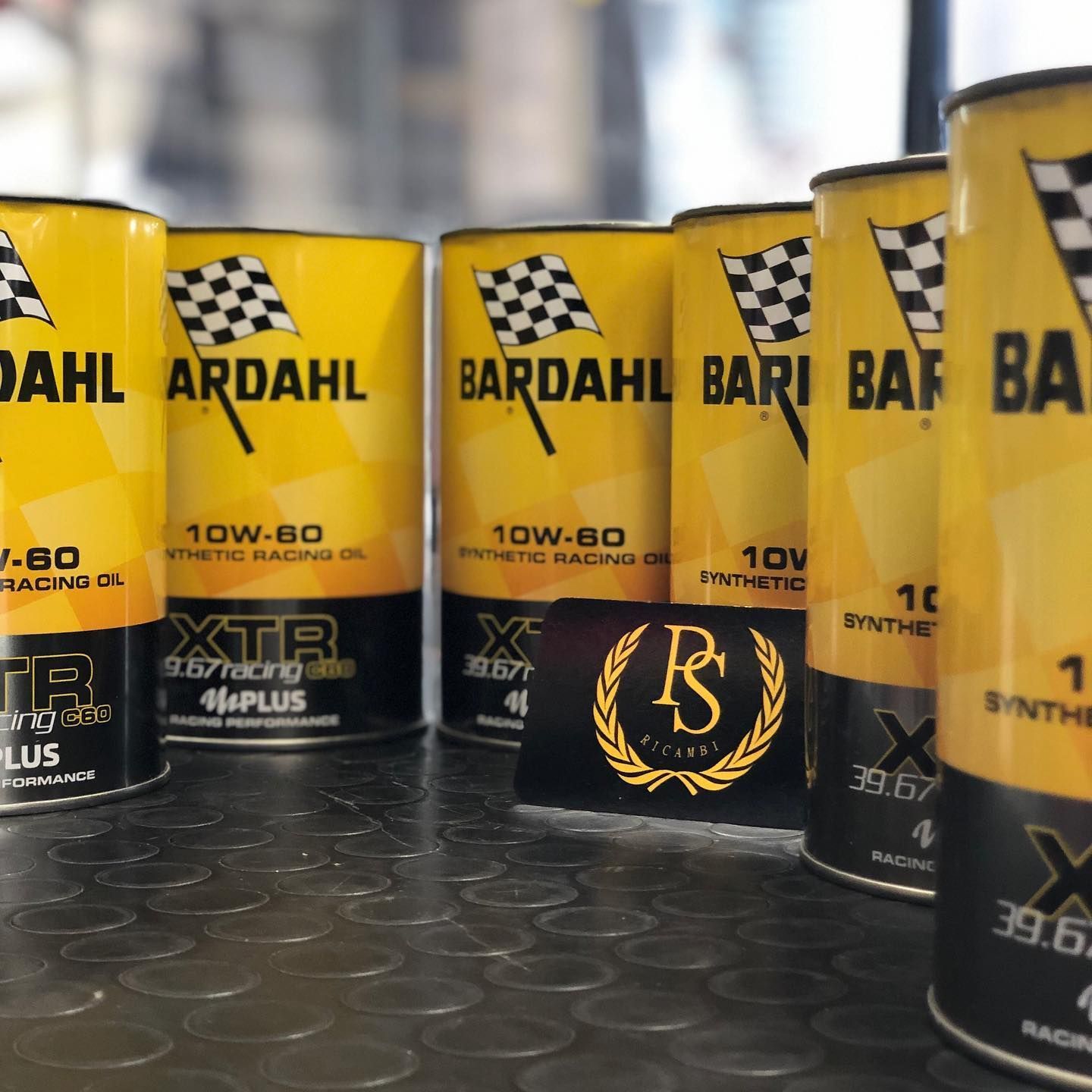 Una fila di lattine gialle di olio sintetico da competizione Bardahl XTR 10W-60 esposte su una superficie scura e ruvida, con un cartellino con il logo.