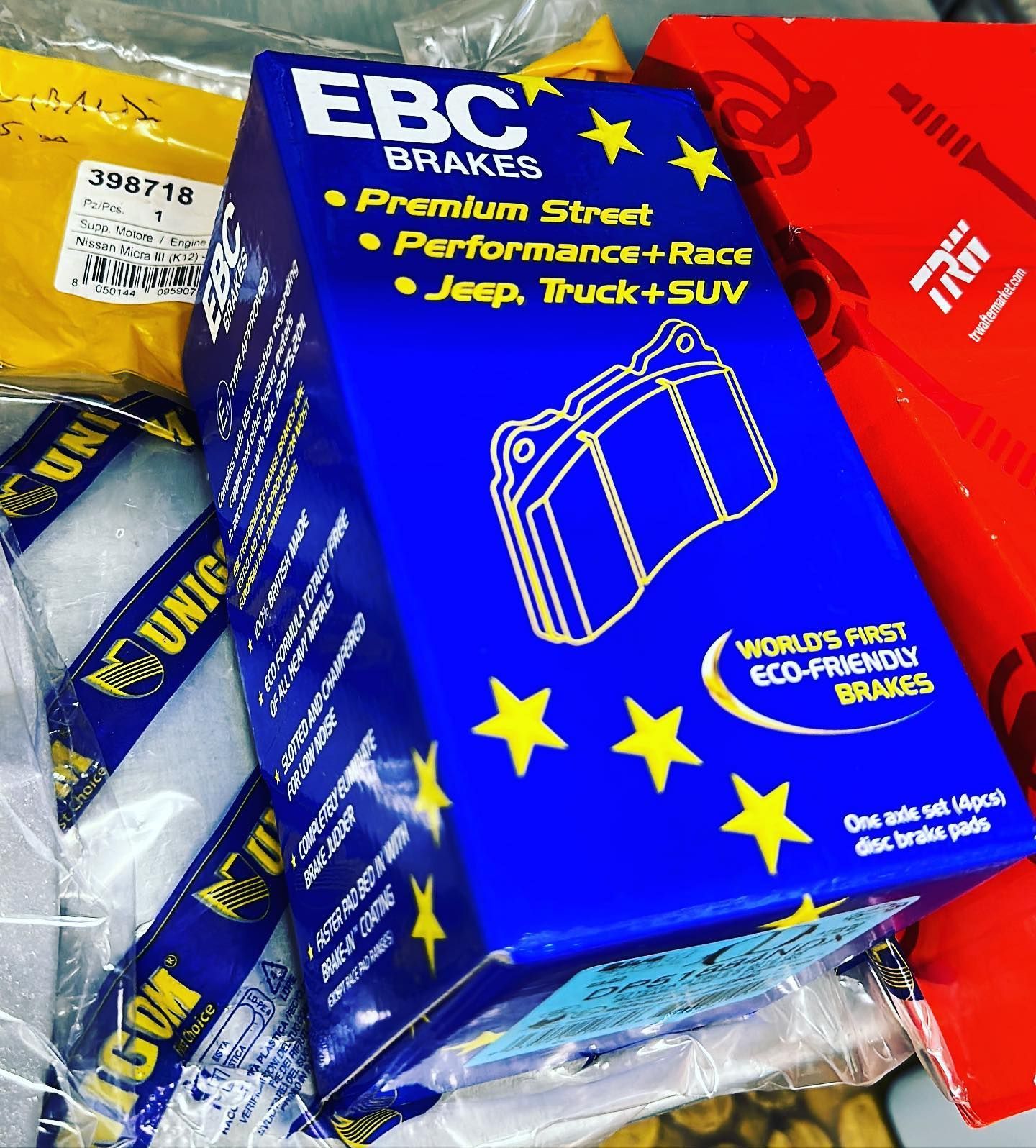 Una scatola blu EBC Brakes per pastiglie freno a disco per Jeep, camion e SUV, circondata da altri imballaggi di ricambi auto.