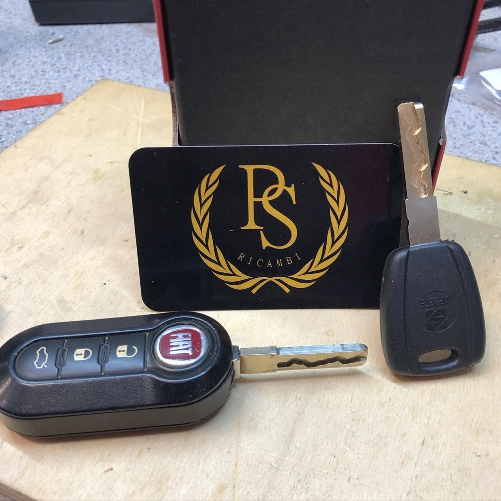 Un portachiavi a scatto nero della Fiat e una chiave di riserva posizionati davanti a un cartoncino nero con il logo PS dorato e una corona d'alloro.