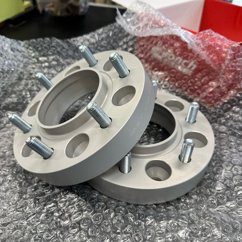 Due distanziali per ruote in metallo grigio con perni filettati, appoggiati su pluriball, con una scatola rossa sullo sfondo.