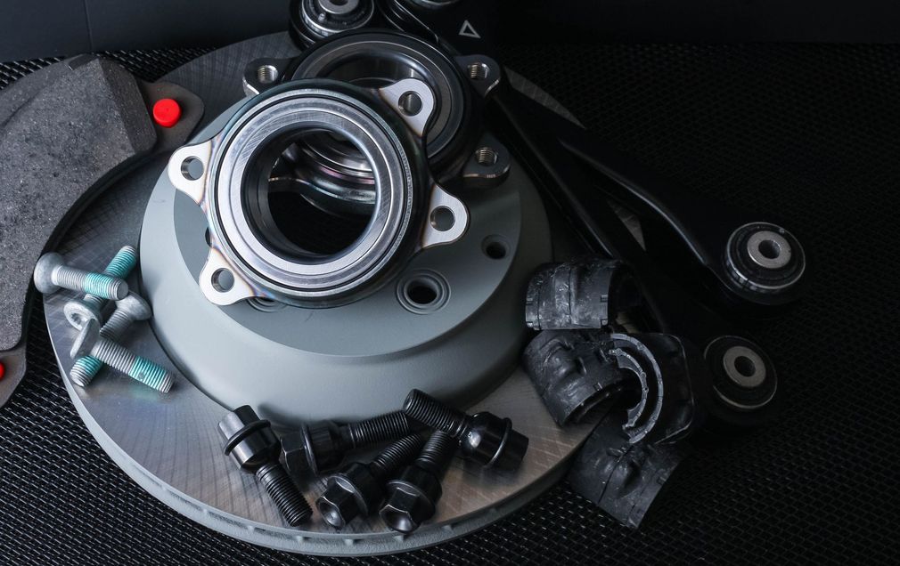 Nuovi componenti delle sospensioni per auto, tra cui un disco freno, un cuscinetto ruota, un braccio oscillante e bulloni di fissaggio, su sfondo scuro.