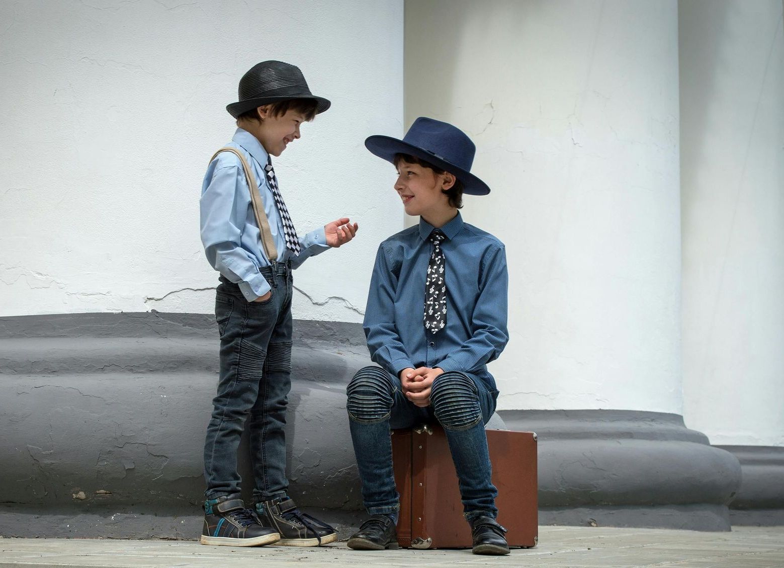 Two young boys are sitting on a suitcase and talking to each other.