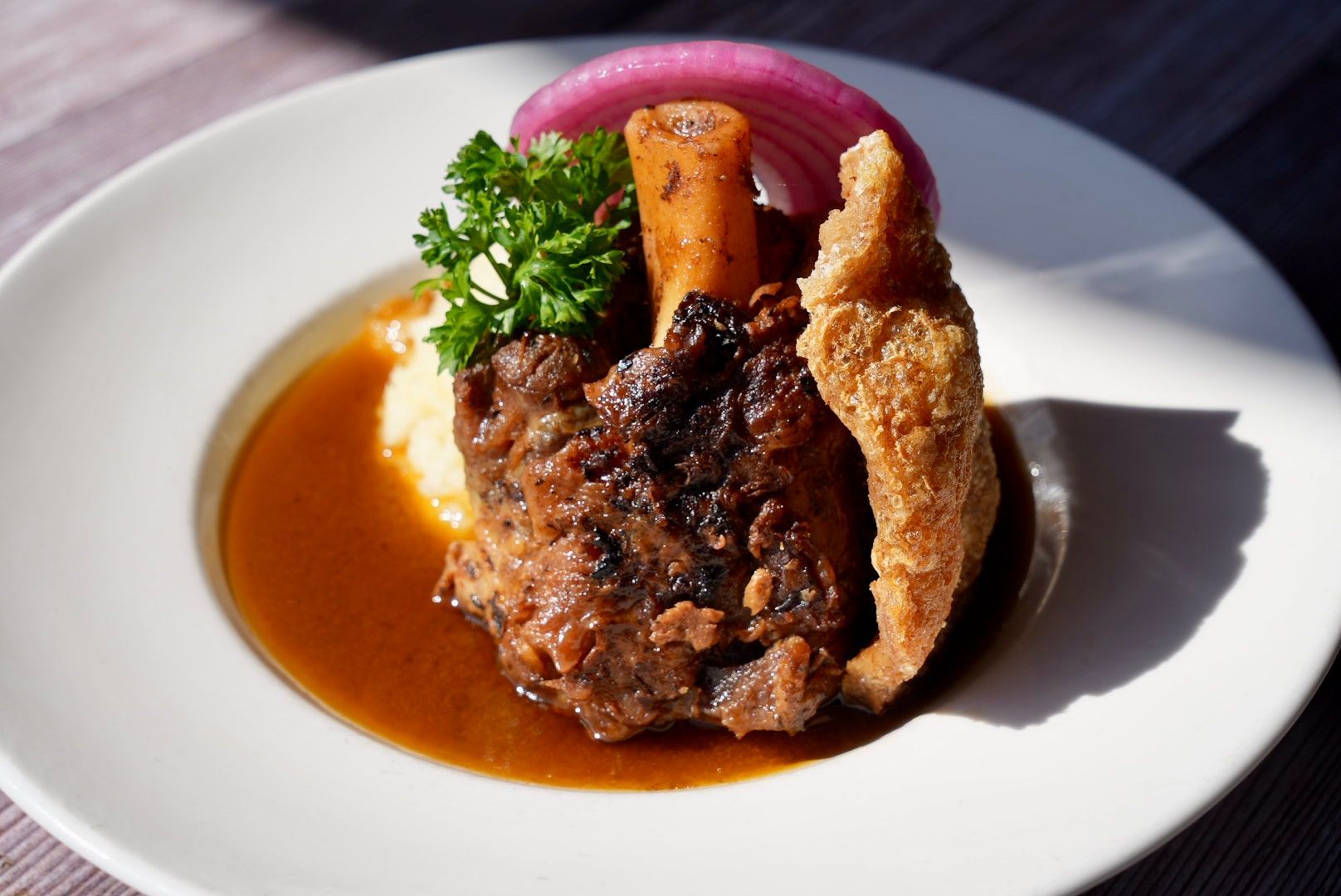 Braised meat shank, gravy, polenta, and fried pork rind on a white plate; garnished with red onion and parsley.