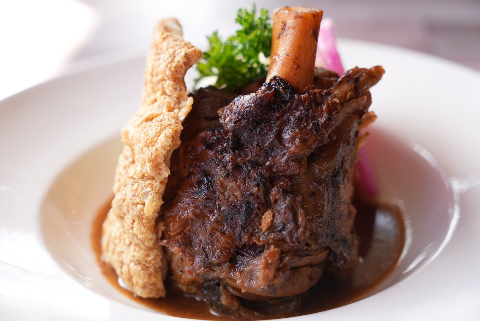 Braised lamb shank on a white plate with sauce, bone protruding, accompanied by a fried side and garnish.