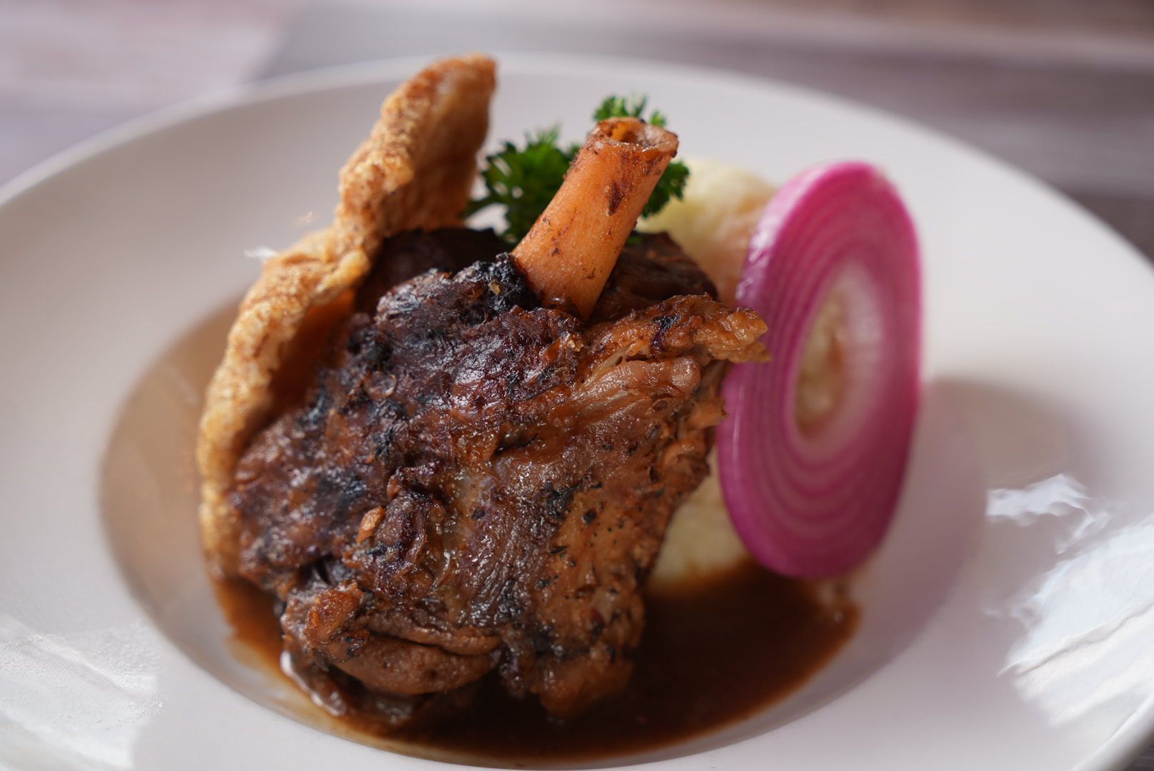 Braised lamb shank with mashed potatoes, red onion, and crispy pastry on a white plate.