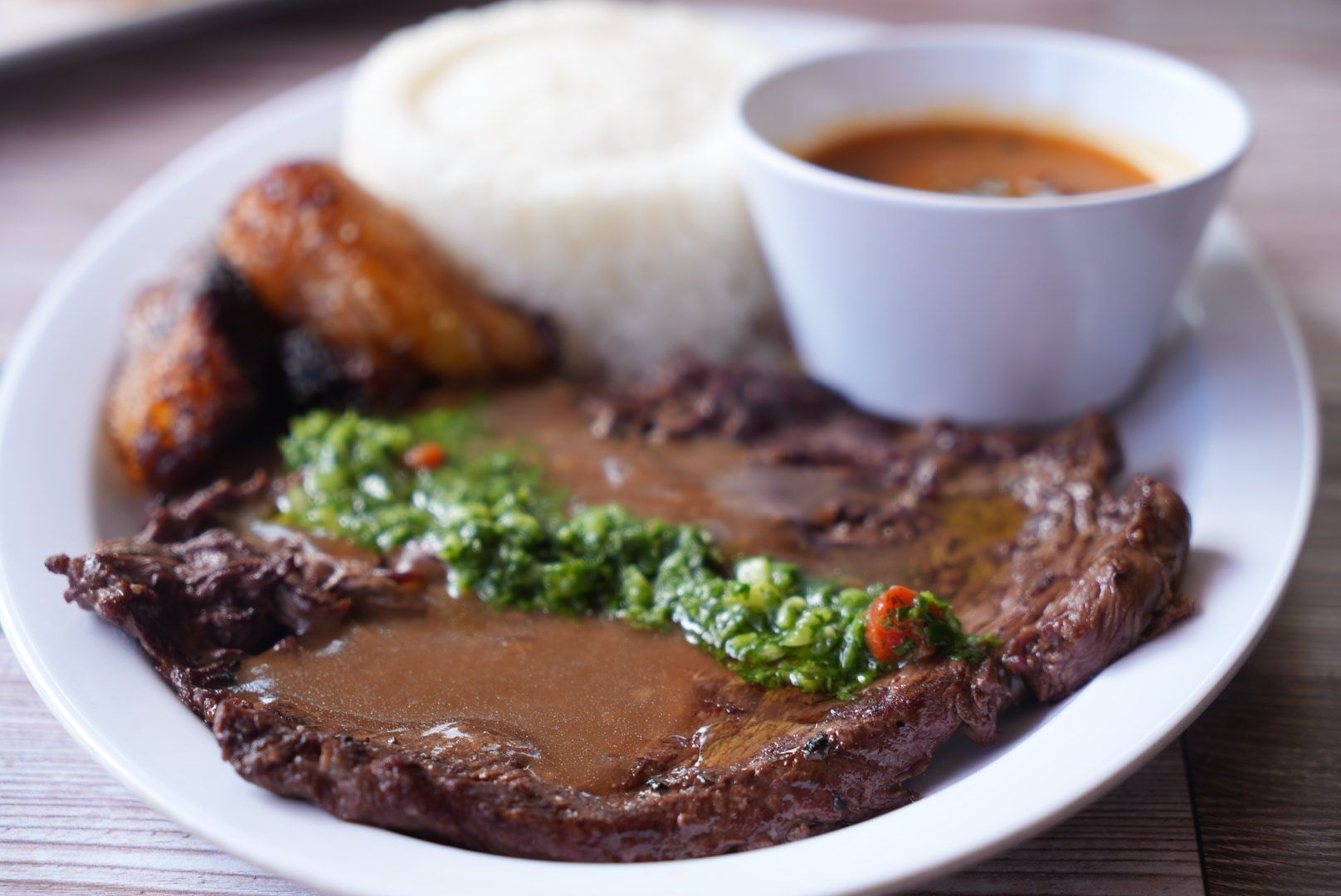 Plate of food: steak with green sauce, rice, fried plantains, and a side of sauce.