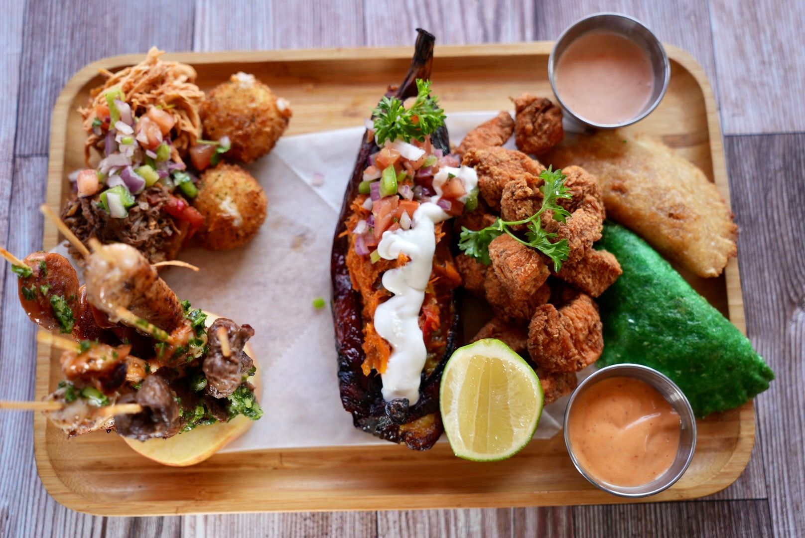 Wooden tray with various fried foods: eggplant, chicken, skewers, balls, sauces, lime, and a green pastry.