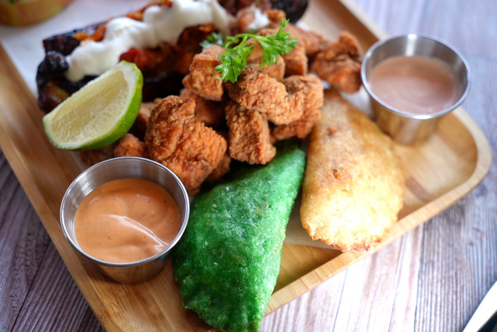 Wooden platter with fried chicken, green tortilla, fried pastry, lime wedge, and dipping sauces.