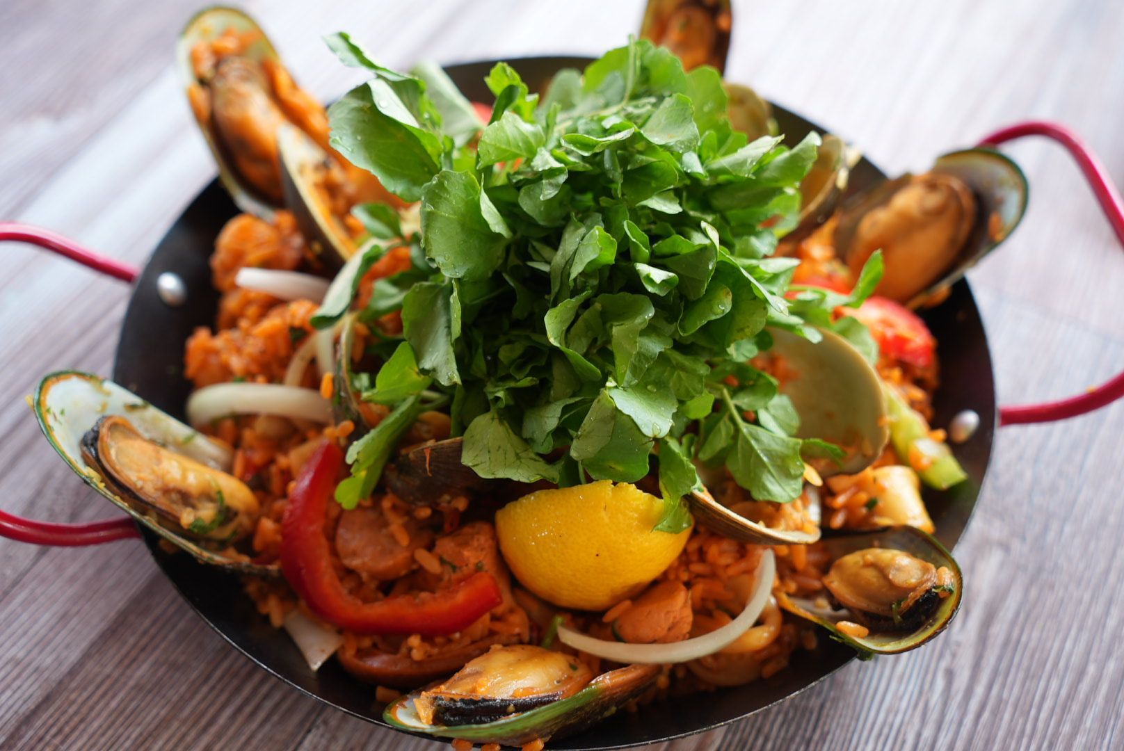 Paella in black pan, mussels, greens, lemon. Red peppers and onions, light wood table.