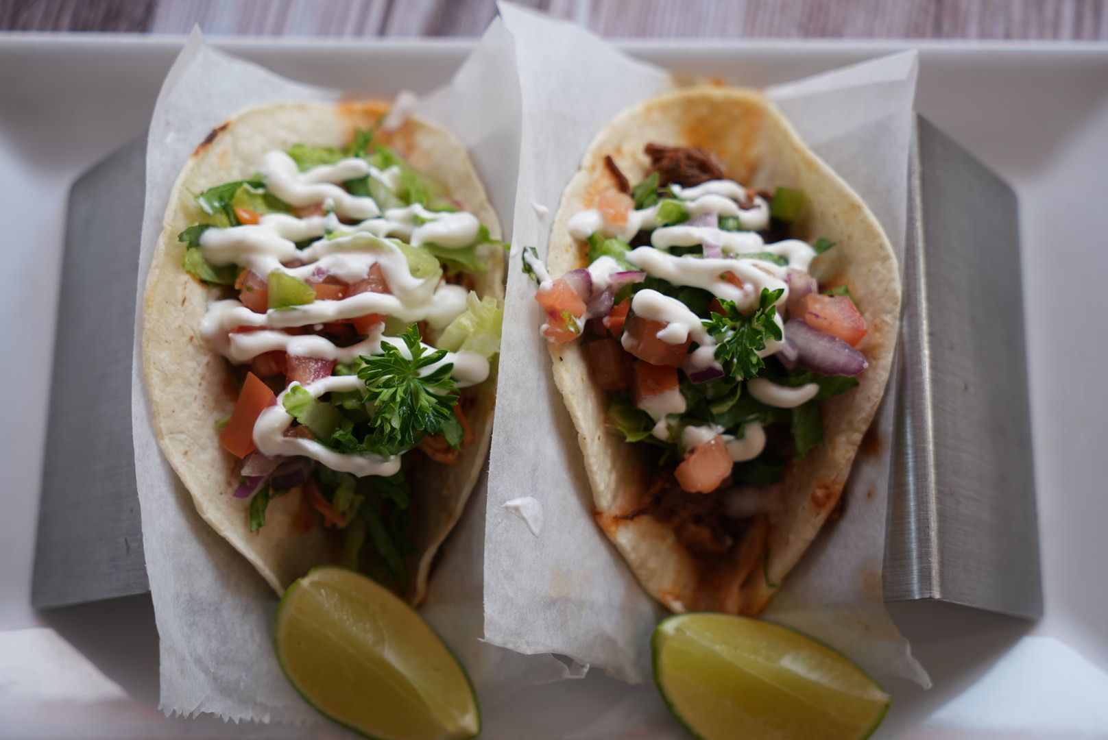 Two tacos with toppings, drizzled with white sauce, on a white plate with lime wedges.