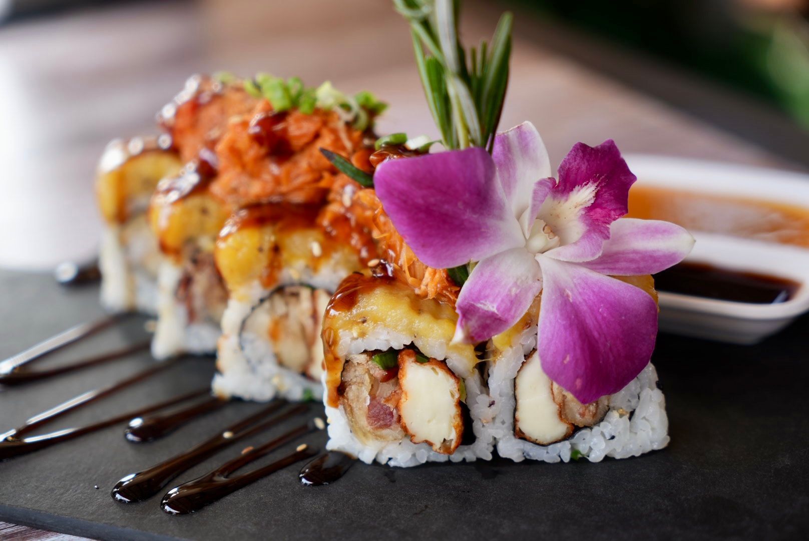 Sushi roll topped with crab, sauce, and orchid flower, on a slate with soy sauce.