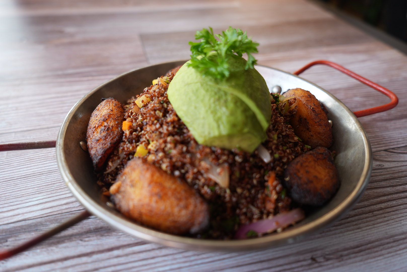 Dish of quinoa, plantains, avocado, and parsley in a metal bowl on a wood table.