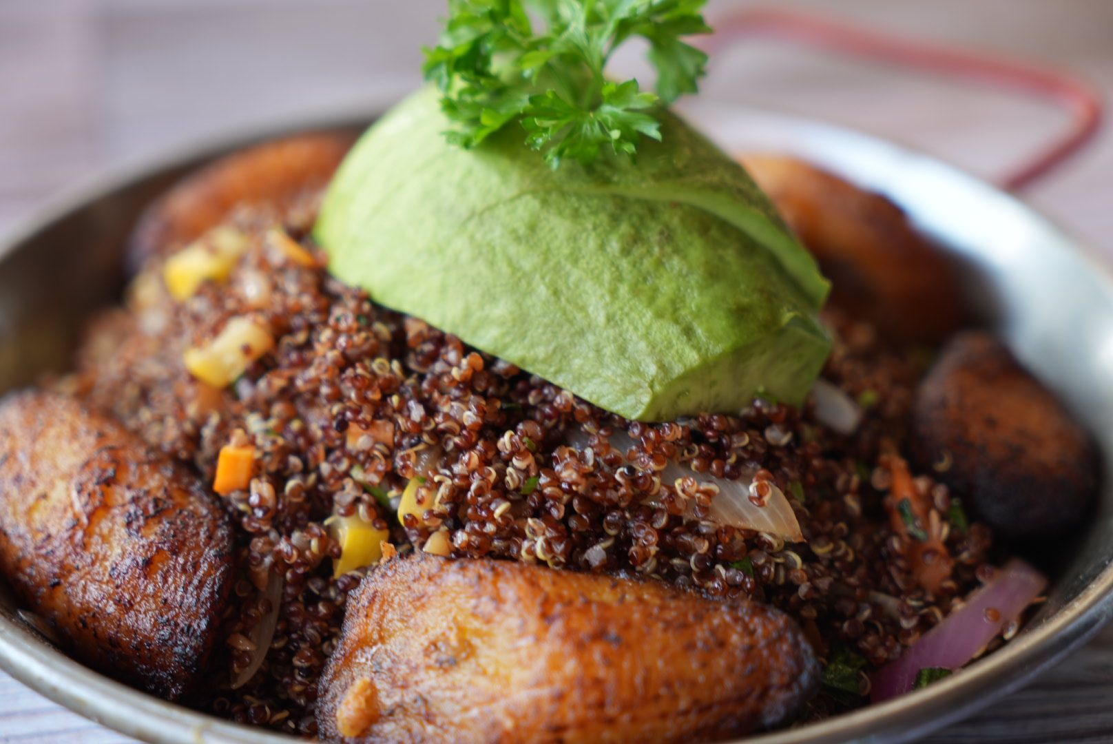Quinoa bowl with fried plantains, avocado slice, and parsley garnish in a metal bowl.