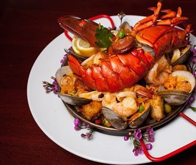 Seafood paella in a pan on a white plate; lobster, clams, shrimp, and lemon garnish.