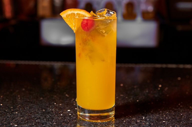 Tall glass of orange cocktail with ice, garnish of orange slice and cherry on a bar.