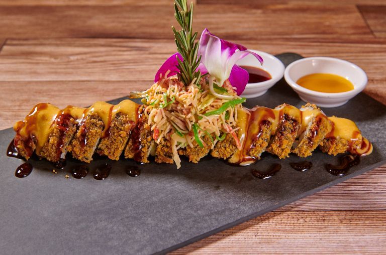 Fried sushi roll with crab salad garnish, sauces, and floral arrangement on a dark platter.