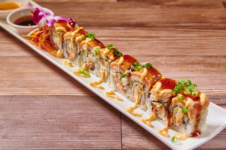 Sushi roll topped with sauce, arranged on a white rectangular plate with dipping sauce and garnish.