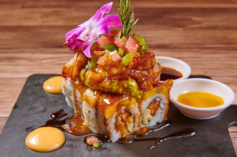 Sushi roll with toppings, garnished with a flower and sauce on a dark serving board.