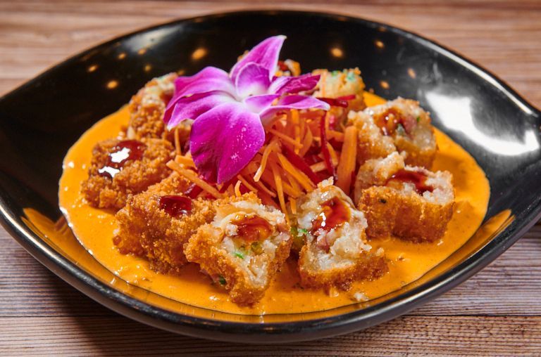 Fried sushi rolls with orange sauce, garnished with shredded carrots and a purple flower, in a black bowl.
