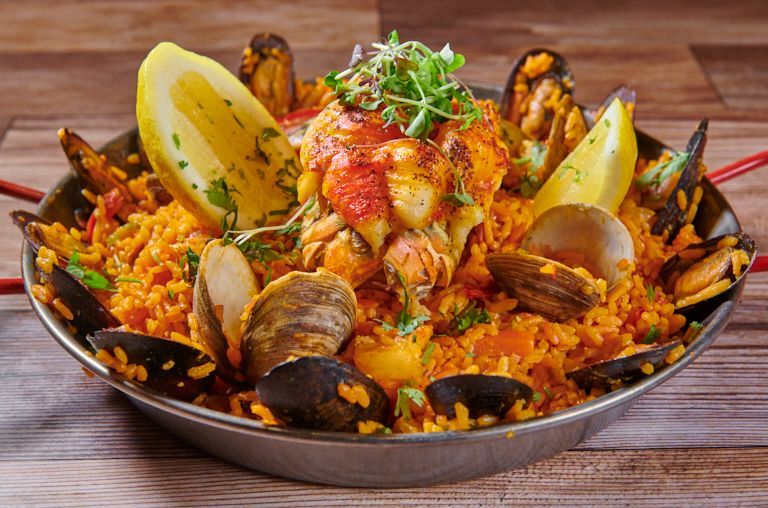 Seafood paella in a pan, featuring mussels, clams, lobster, and lemon wedges, atop vibrant yellow rice.