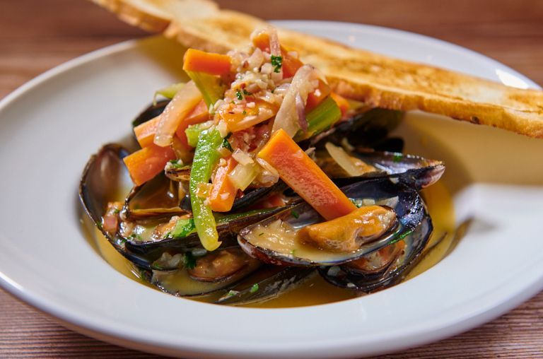 Mussels in broth with vegetables and toasted bread in a white bowl.