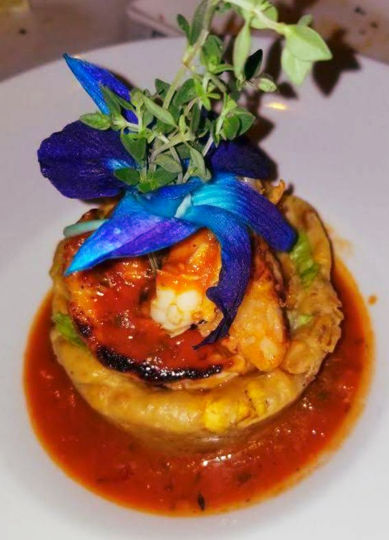 Shrimp dish topped with blue flowers and herbs, served in tomato sauce.