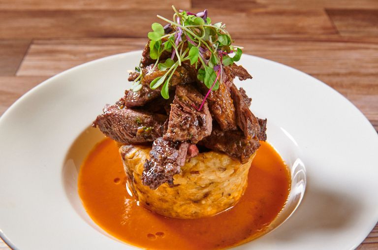 Steak dish: sliced meat atop a potato cake, surrounded by red sauce, garnished with sprouts, on a white plate.