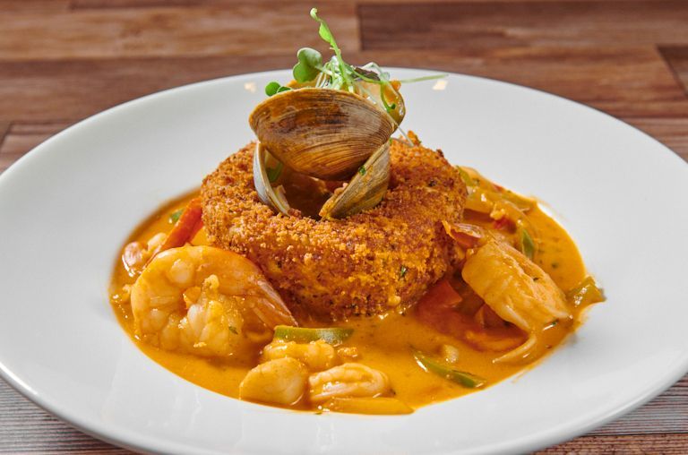Seafood dish on a white plate: fried patty topped with clams, surrounded by shrimp and creamy orange sauce.