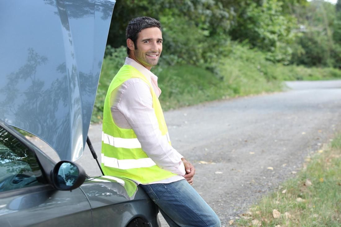 A man in a yellow vest is leaning against a broken down car.