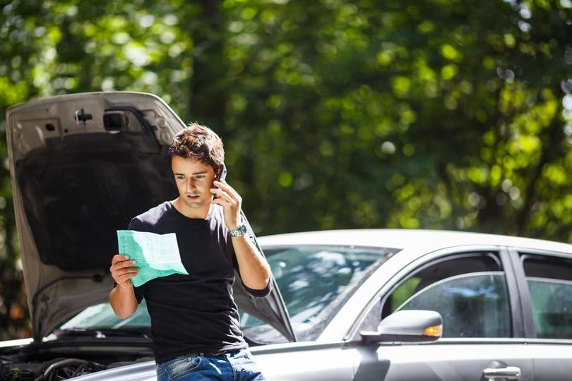 A man is standing next to a broken down car talking on a cell phone.