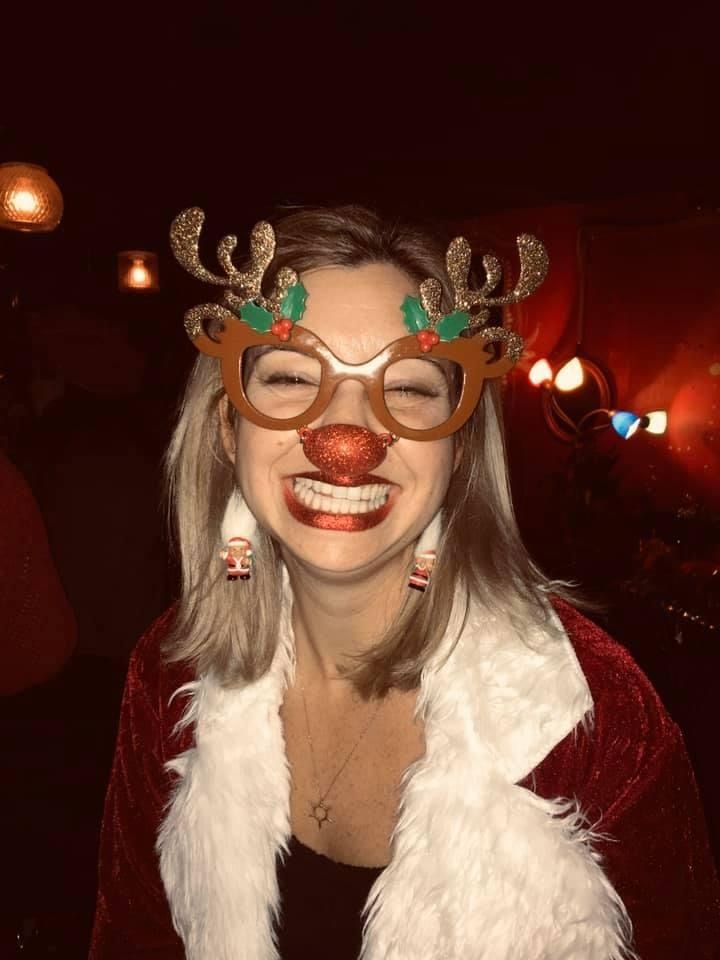 A woman wearing a santa claus costume and reindeer glasses is smiling.
