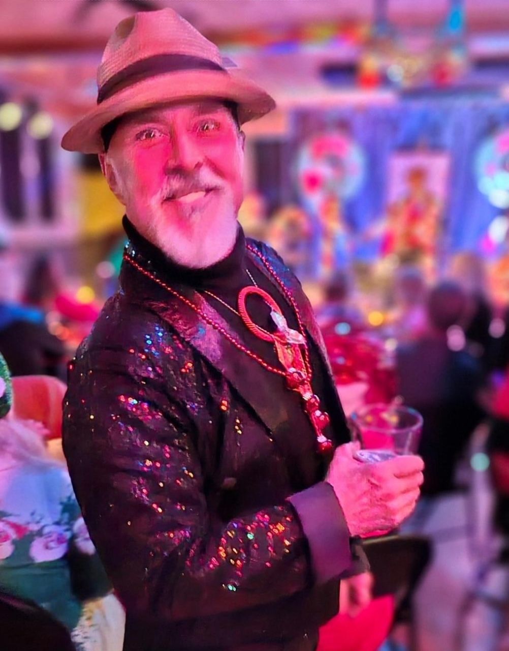 A man wearing a hat and sequined jacket is holding a glass of wine.